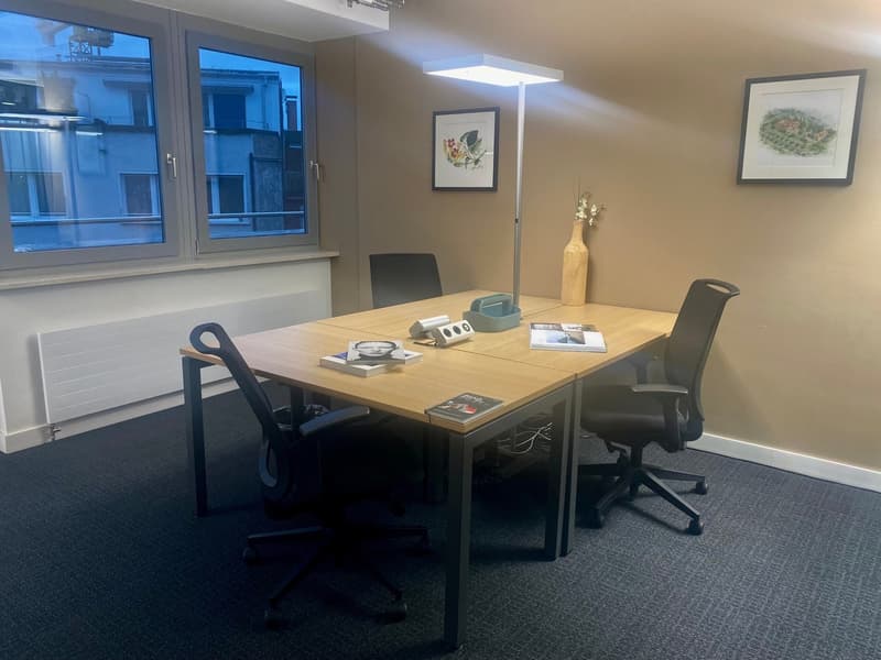 A room with a long wooden table, four black swivel chairs, a white radiator, a floor lamp, a vase with flowers, and two framed pictures on the wall.