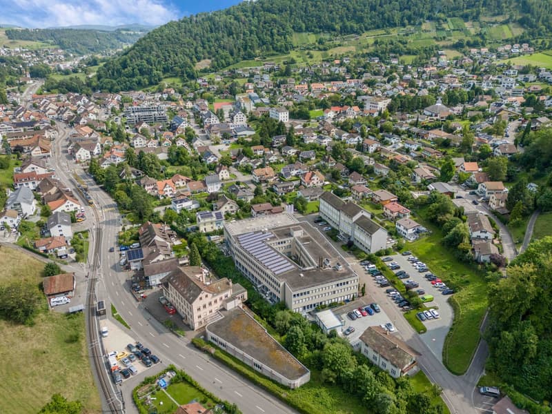 An aerial view of a building with multiple structures including residential houses and buildings, surrounded by greenery and several cars parked.