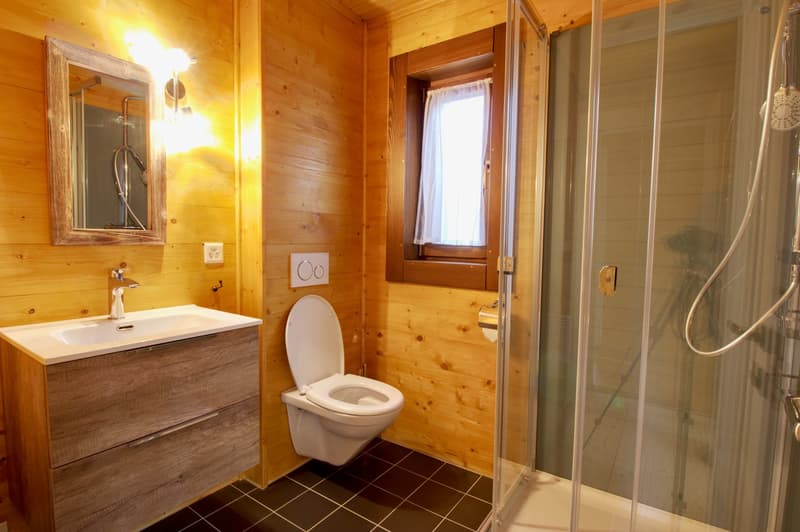 Bathroom with wooden walls, white toilet, sink with mirror, shower enclosure, and window with white curtains.