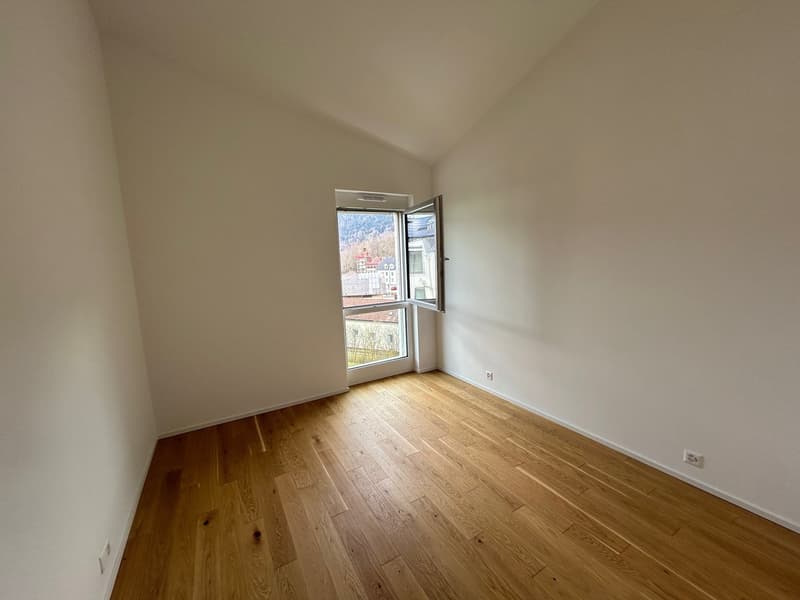 The image shows a spacious bedroom with a large window overlooking a scenic view. The room has white walls and a hardwood floor, creating a clean and minimalist aesthetic. The window allows natural light to flood the space, making it feel bright and airy.