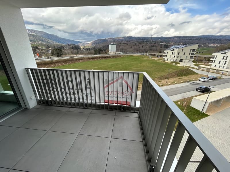 Balcony with a view of a grassy field, surrounded by apartment buildings and mountains in the background