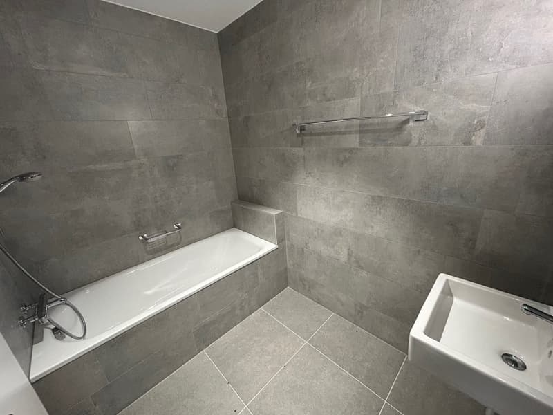 Large bathroom with grey tile walls and floor, a white bathtub, and a white sink.