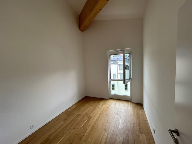 This image shows an empty room with white walls, a wooden ceiling beam, and a large window that provides natural light. The room has a hardwood floor and appears to be a living or bedroom space.