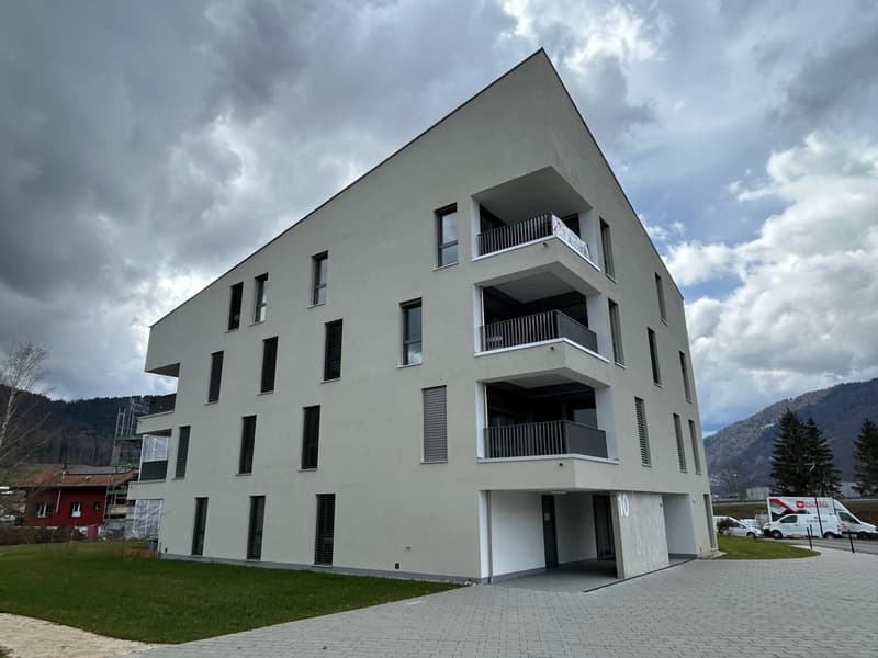 Multi-story apartment building with a modern, minimalist design. The building has a gray exterior with large windows and balconies on each floor. There is a paved walkway and landscaping in the front of the building.