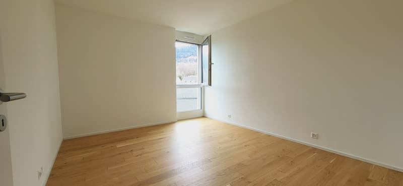 Spacious bedroom with hardwood flooring, white walls, and a large window providing natural light and a view of the outdoors.
