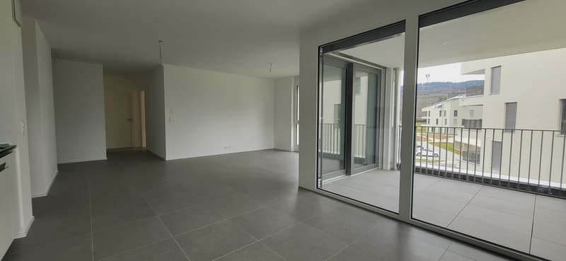 Spacious open-plan living room with large sliding glass doors leading to a balcony, grey tile flooring, and neutral wall colors.