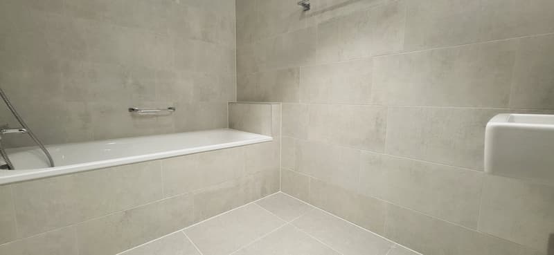 Large bathroom with gray tile walls and floor, a white bathtub, and a shower fixture.