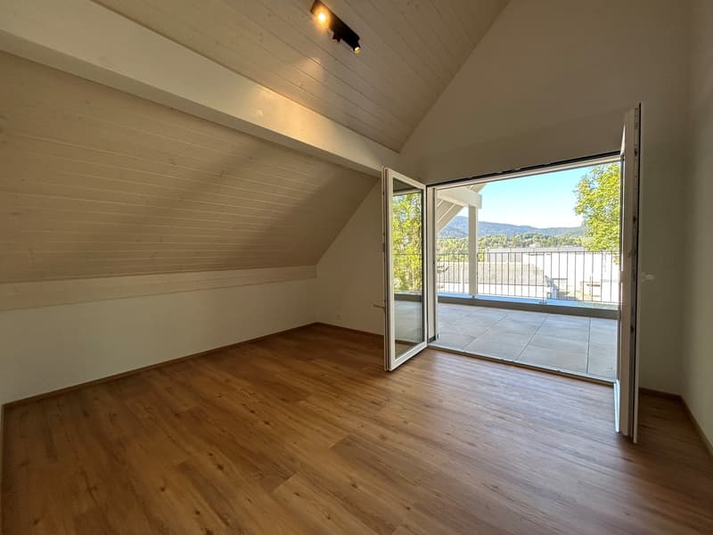 open-plan attic room with wooden floors, ceiling with beams, large windows leading to a balcony, no furniture