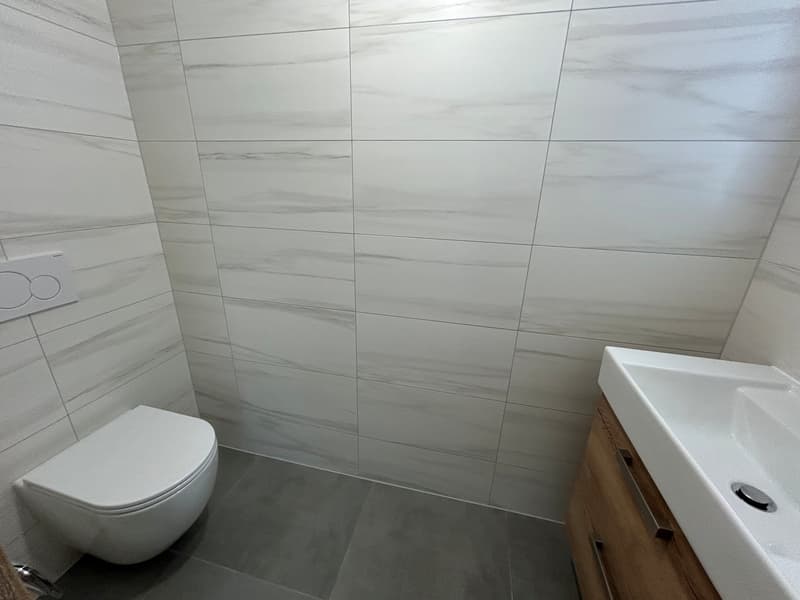 Modern bathroom with white tiles, white toilet, gray tiles on the floor, wooden cabinet under the sink, white sink