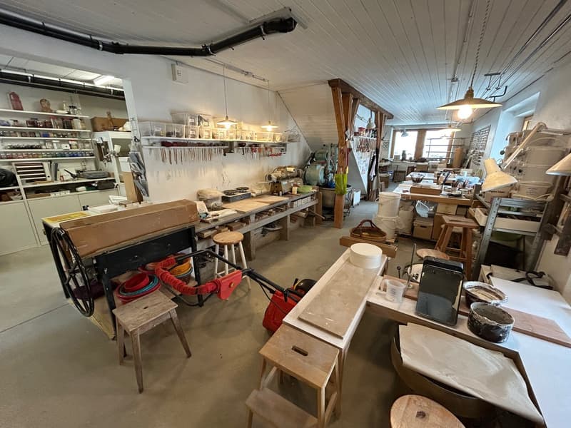 spacious workshop with various wooden tables, workbenches, and storage shelves, hanging lamps, stools, equipment, buckets, and barrels, wooden beam supports, and ceiling lamps.