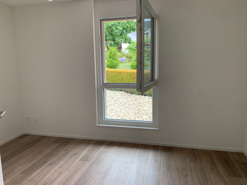 Empty room with wooden floor, white walls, and a large window with a view of a garden.