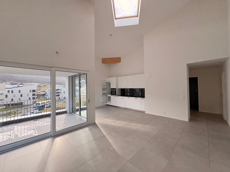 Bright, empty room with white walls and ceiling, large grey tiles on the floor, skylight, sliding glass doors to a balcony, white kitchen with modern appliances, white cabinets.