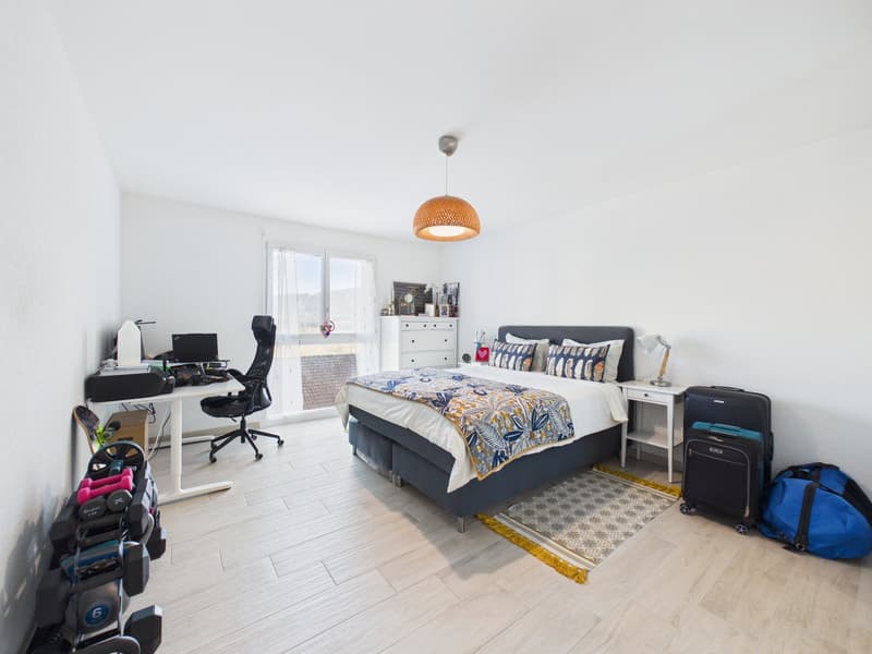 Spacious bedroom with bed, white walls, wooden floor, large window, desk, chair, wardrobe, suitcases, rug, lamps, pillows, and bedspread.