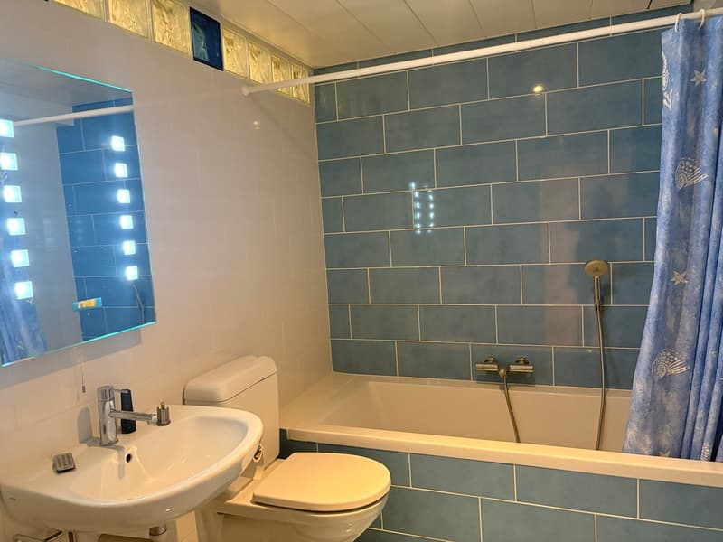 blue tiles, mirror with lighting, sink, toilet, bathtub with shower, blue curtain