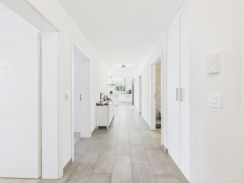 Empty hallway with white walls, tiled floor, white doors, white switchboards, white cabinet, and a chandelier