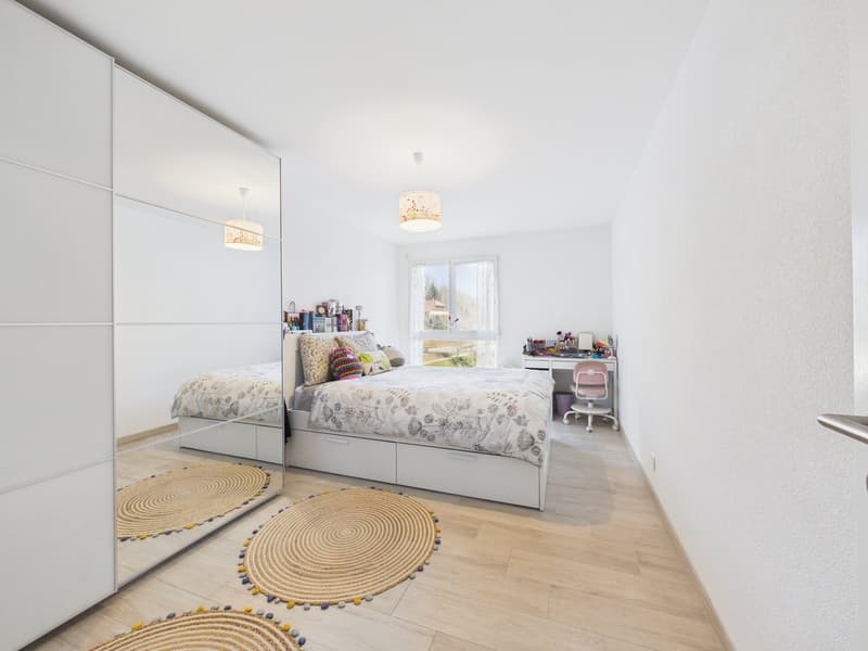 Bedroom with a bed, desk, chair, and a large mirror. The room has wooden floors and white walls. There are two round rugs on the floor.