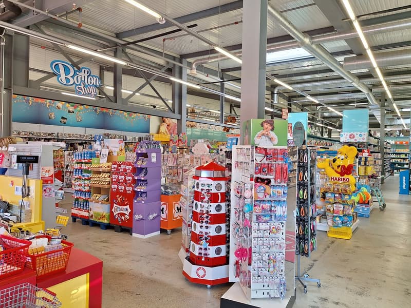 The image shows the interior of a retail store, likely a convenience or variety store. The store has a large open floor plan with various shelves, displays, and racks filled with a wide variety of products. The store has a bright and colorful aesthetic, w