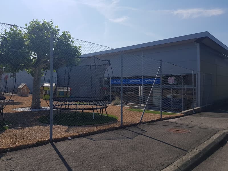 The image shows an outdoor recreational area with a large covered structure, a trampoline, and a small playhouse. The area is surrounded by a wire mesh fence and has a paved surface with some grassy areas. There appears to be a parking space or area nearb