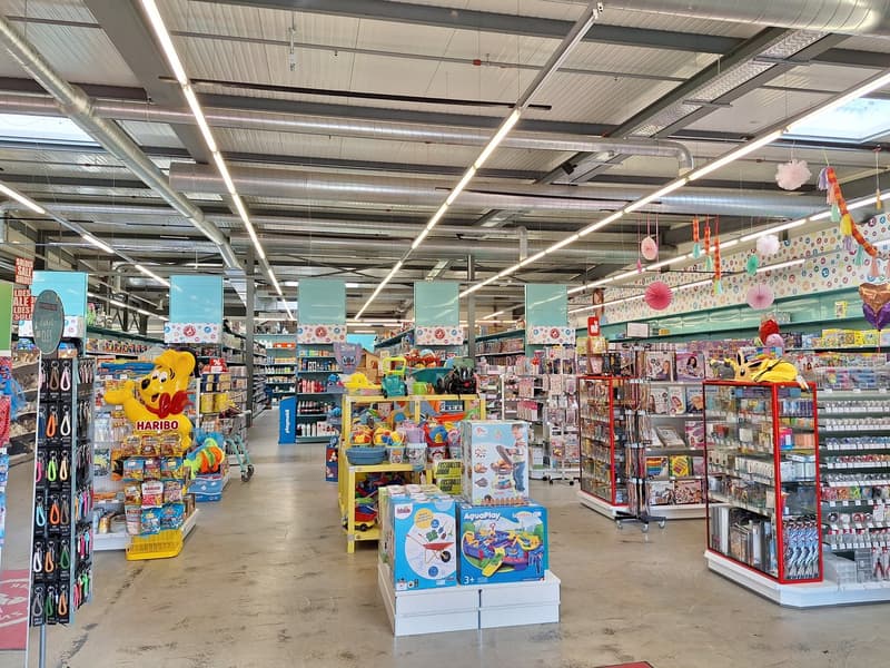 The image shows the interior of a retail store, likely a toy or children's store. The store is filled with various colorful toys, games, and other merchandise displayed on shelves and racks. The ceiling has exposed ductwork and lighting fixtures, creating