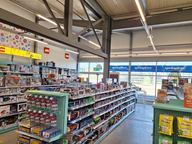 The image shows the interior of a retail store, likely a toy or hobby store. The store has high ceilings with exposed beams and racks filled with various products, including Lego sets and other toys. The store has large windows that provide natural light
