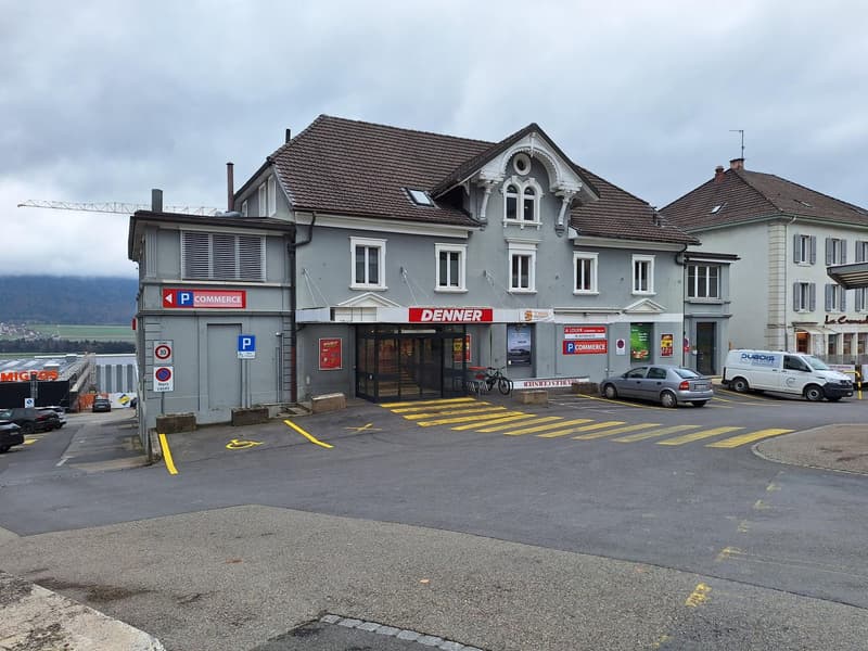 2-story commercial building with a Denner store on the ground floor, parking spaces in front, and residential units on the upper floors. The building has a traditional architectural style with a sloped roof and decorative elements.