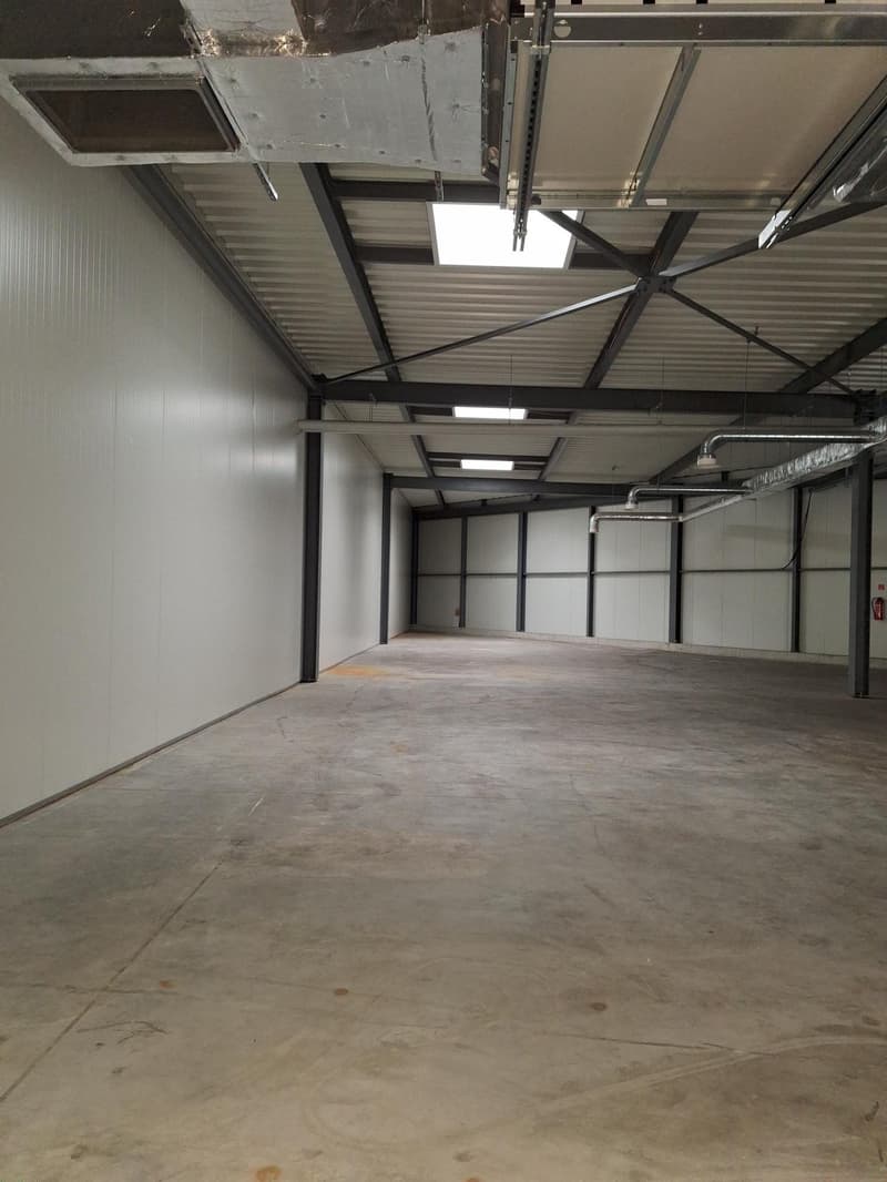 The image shows a large, open warehouse space with a concrete floor, metal framing, and corrugated metal roofing. The space appears to be empty and unfinished, with exposed pipes and electrical conduits visible overhead.