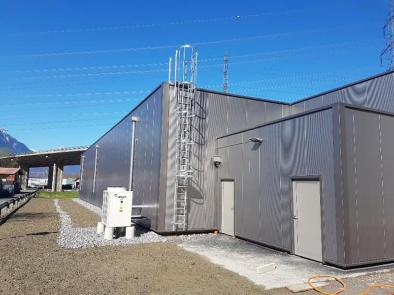 Large gray industrial building with a ladder on the side, surrounded by a gravel parking area.