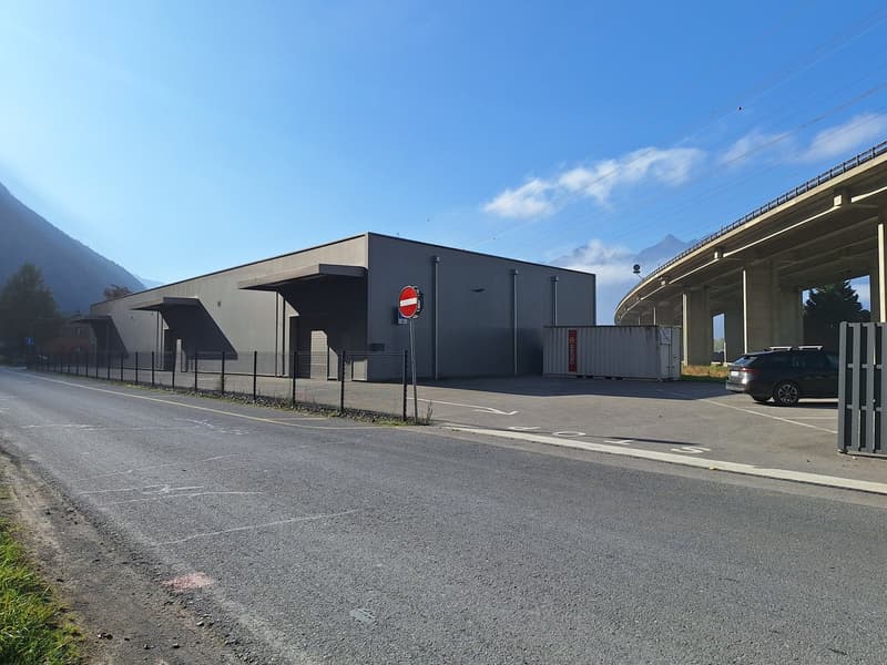 A large, modern commercial building with a dark exterior and a parking area in front. The building appears to be situated in a mountainous area with a clear blue sky in the background.