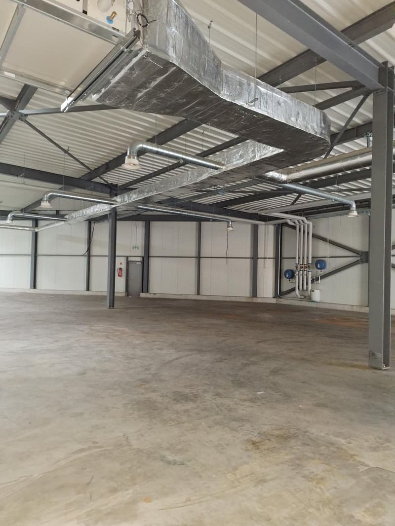 Large open warehouse space with concrete flooring, metal beams and ductwork overhead, and storage units or equipment visible.