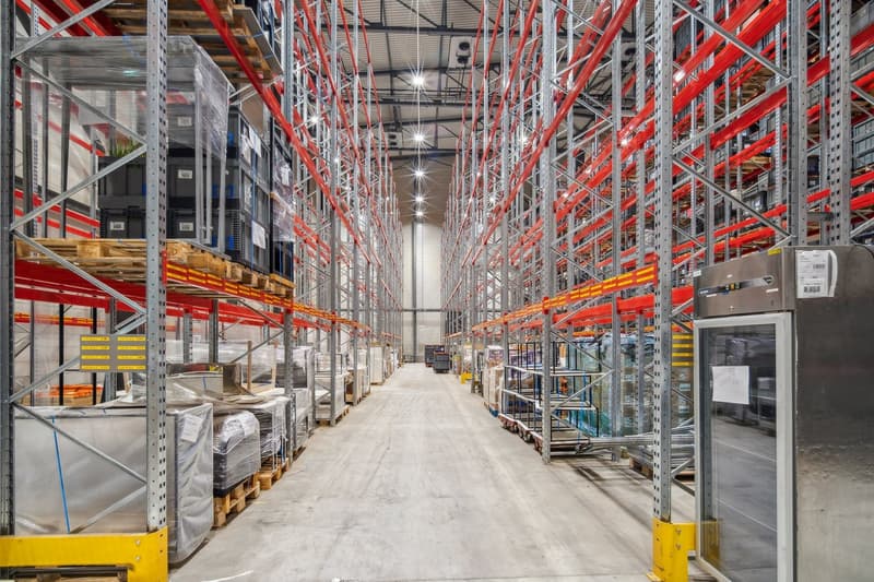 Large warehouse with metal shelving units, storage boxes, and pallets, concrete floor, metal beams, and lights