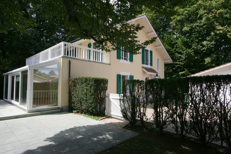 yellow house with green shutters, glass windows, tiled driveway, glass terrace, hedges
