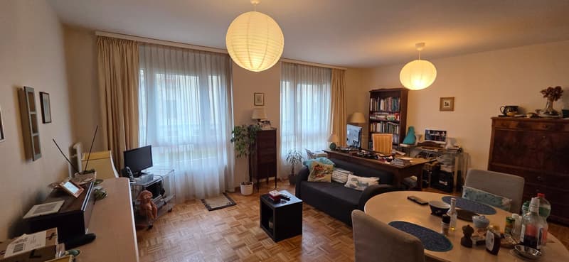large open living space with hardwood floors, windows with white curtains, a black couch with pillows, a brown wooden desk, a round table, round table lamps, framed picture, and a potted plant.