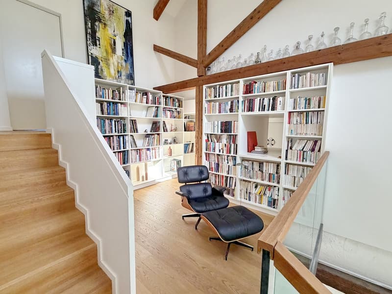 room with wooden floors, white walls, wooden beams, wooden staircase, bookcases, glass handrail, chair, ottoman