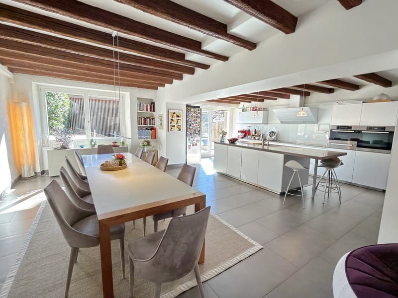 large kitchen, dining area, wooden ceiling, white tiles, long dining table, gray chairs, countertop island, bar stools, white cabinets, refrigerator, microwave, oven, picture frames, bookshelf, window