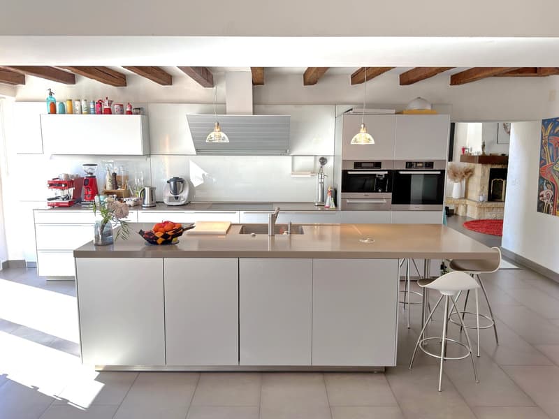 Modern white kitchen with island, undermount sink, central gas stove, built-in microwave, refrigerator, two ovens, and dishwasher.