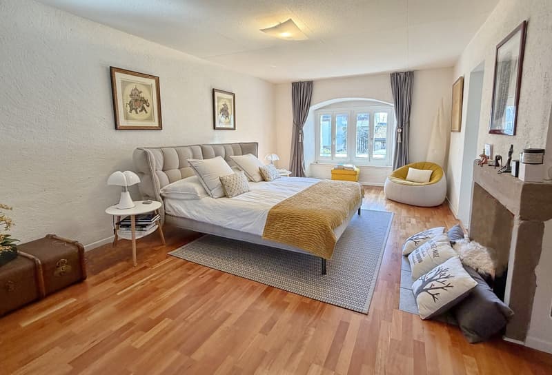 modern bedroom with large bed, white walls, gray rug, bedside table, decorative pillows, fireplace, arched window, chair, lamp