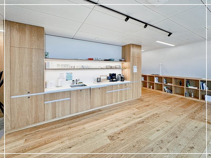 The image shows a modern office space with a kitchenette area. The space features wooden cabinetry and shelving, a counter with a sink and what appears to be a dishwasher, and a bookshelf area along the back wall. The flooring is a warm-toned hardwood, an