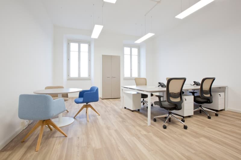 Bright and modern office space with large windows, white walls, hardwood floors, a round table with chairs, a desk with office chairs, and a storage cabinet.