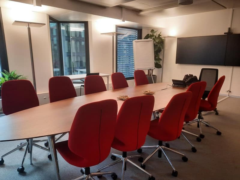 Long wooden conference table, red office chairs, flip chart, white board, ceiling lamps, office plants