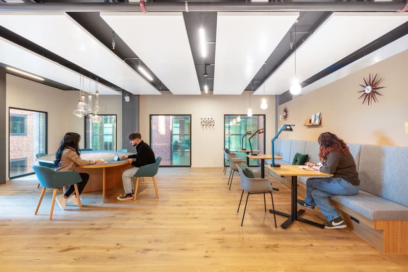 Open area, oval wooden table with chairs, round benches along wall, ceiling lamps, windows, lamps on tables, wooden floor