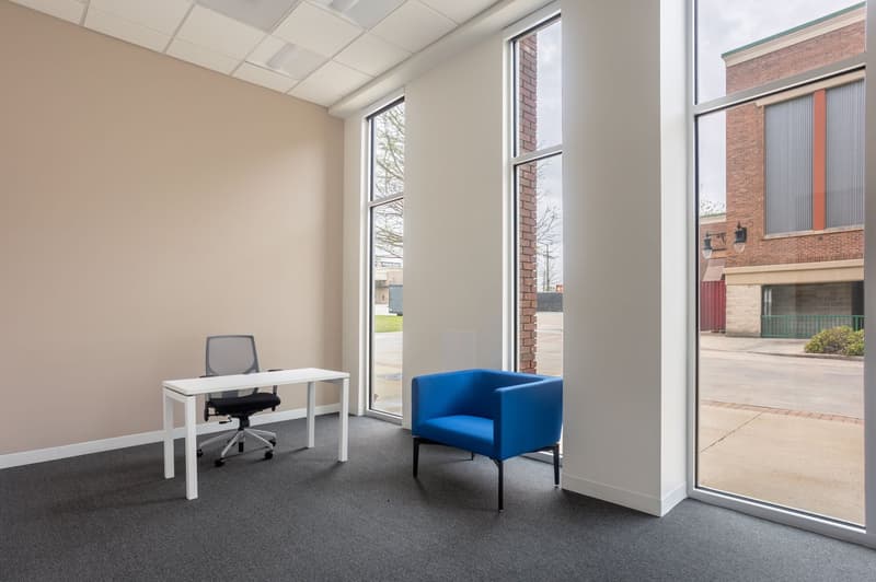 The image shows a modern office space with a white desk, a blue armchair, and large windows overlooking the exterior. The room has a neutral color scheme with beige walls and gray carpeting. The overall design appears to be minimalist and professional.
