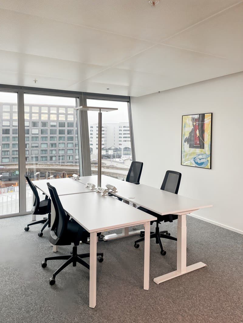 This image shows a modern office space with large windows providing a view of the surrounding buildings. The room has a minimalist design with white walls, a gray carpet floor, and a long rectangular table with black office chairs. There is also a framed