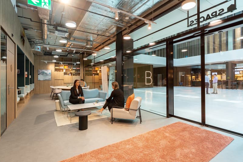 Open office space with modern furniture, large windows, and exposed ceiling. Two people are seated on couches having a conversation.
