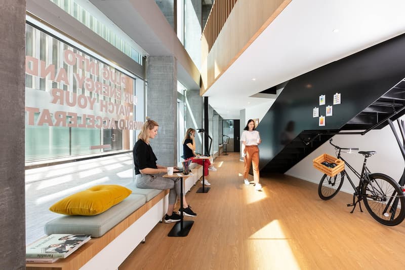 Modern and open office space with large windows, wooden floors, comfortable seating areas, and a bicycle parked inside. The space has a relaxed and collaborative atmosphere.