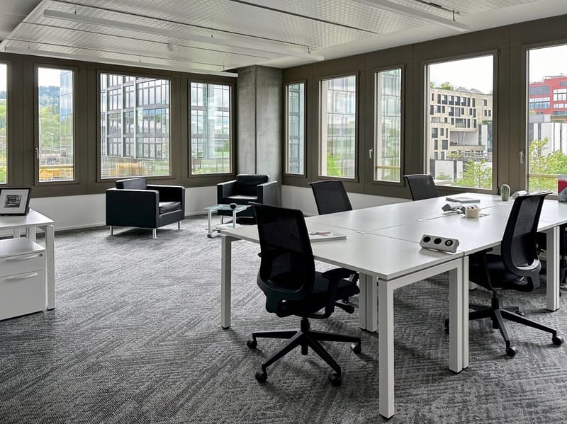 Large open office space with floor-to-ceiling windows providing a view of the surrounding buildings. The space features a conference table with chairs, as well as individual workstations with desks and chairs. The walls are dark-colored, and the flooring
