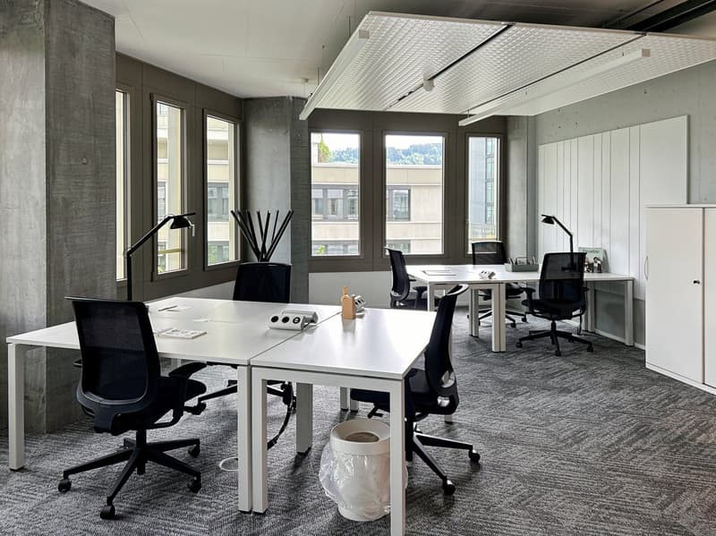 The image shows a modern and minimalist office space. The room has large windows providing natural light, concrete walls, and a high ceiling with a grid-like lighting fixture. The office is furnished with white desks, black office chairs, and various offi