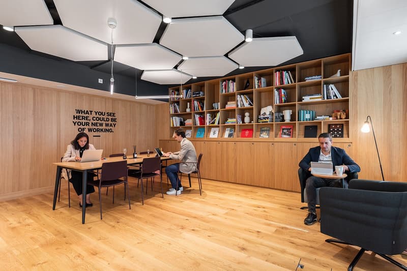 modern office space with wooden floors, wooden shelves, large hexagonal ceiling fixtures, two round tables, four chairs, three individuals sitting
