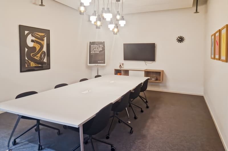 Large conference table with black chairs, modern lighting fixture, wall-mounted TV, shelving unit, and framed artwork on the walls