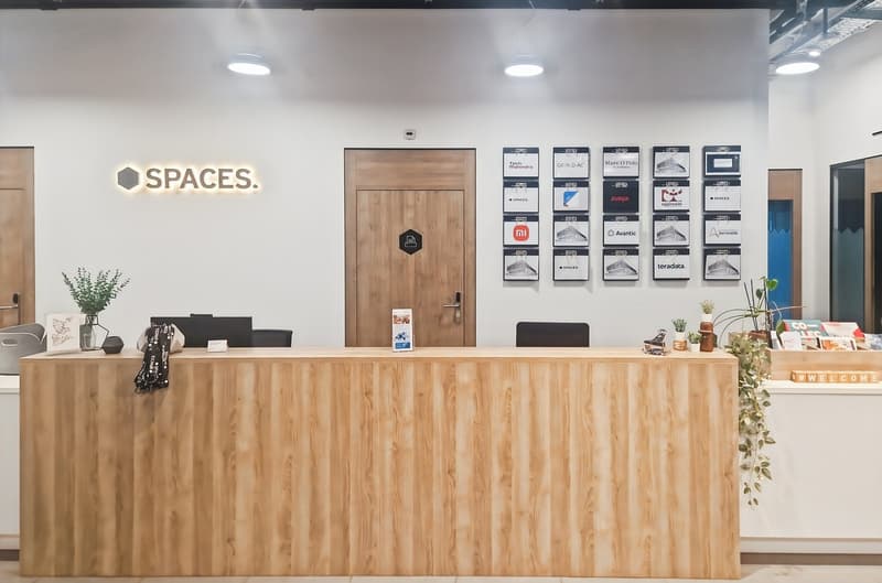 Wooden reception desk, white walls, wooden door, multiple frames with various brand logos, plants, seating area, welcome signage