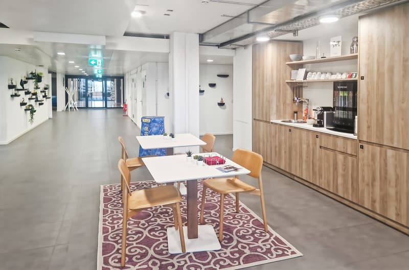 The image shows a modern and stylish office break room or kitchen area. It has a wooden kitchenette unit with a coffee machine and other appliances. There are several small tables with wooden chairs, and a patterned rug on the floor. The room has a bright